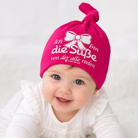 Lachendes Baby trägt pinke Beanie Mütze mit Spruch und Schleifenmotiv.