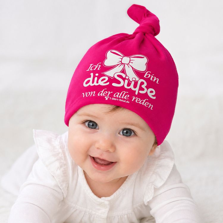 Lachendes Baby trägt pinke Beanie Mütze mit Spruch und Schleifenmotiv.