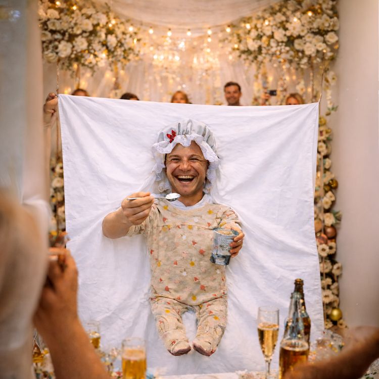 Bräutigam füttern Hochzeitsspiel für Erwachsene & für Geburtstag inkl. Anleitung Mann im Babykostüm lacht während eines Hochzeitsspiels, vor dekorierter Kulisse mit Lichtern und festlicher Atmosphäre.
