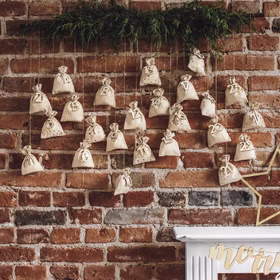DIY Adventskalender Säckchen zum Befüllen 72-teilig mit Holzzahlen SALE Weihnachten DIY Kalender Geschenkidee Advent Adventszeit Rustikale Adventskalender-Dekoration mit nummerierten Stoffbeuteln vor einer Backsteinwand.