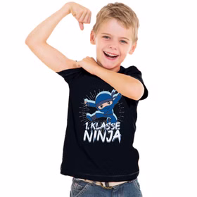 Junge trägt schwarzes Ninja T-Shirt „1. Klasse Ninja“ und zeigt stolz seine Stärke beim Schulanfang.