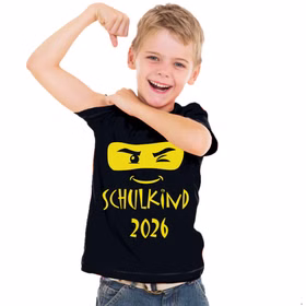 Fröhlicher Junge trägt schwarzes „Schulkind 2026“ T-Shirt mit gelbem Ninja-Motiv und zeigt stolz sein Shirt.