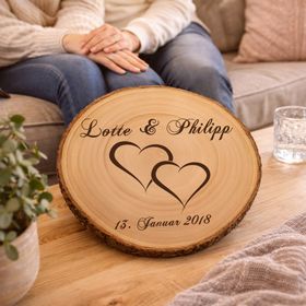 Personalisierte Baumscheibe mit Hochzeitsgravur als Dekoration auf einer festlich gedeckten Hochzeitstafel, im Hintergrund ein Brautpaar und Hochzeitsgäste in feierlicher Atmosphäre.