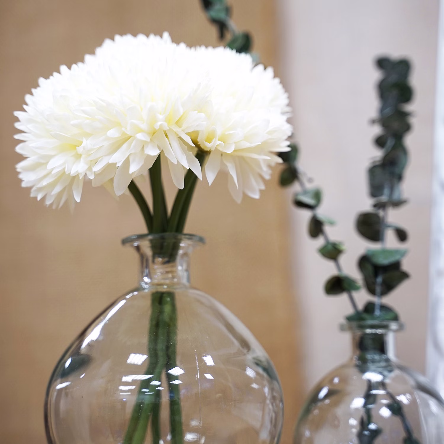 Kunstblumen Dahlien Weiß Glasvase Wohnraumdeko Weiße künstliche Dahlien in einer runden Glasvase als elegante Wohnraumdekoration, daneben eine weitere Vase mit Eukalyptuszweigen.