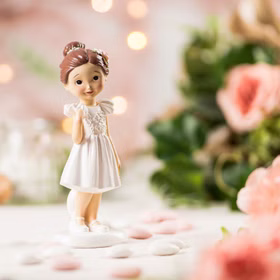 Figur Kommunion Mädchen im weißen Kleid mit goldenem Kreuz und brauner Hochsteckfrisur mit Blüte im Haar von Vorne auf gedecktem Tisch.