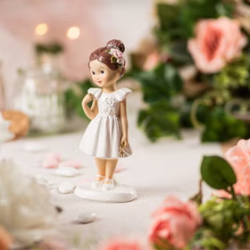 Figur Kommunion Mädchen im weißen Kleid mit goldenem Kreuz und brauner Hochsteckfrisur mit Blüte im Haar von der Seite auf gedecktem Tisch.