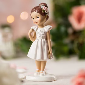Nahaufnahme Figur Kommunion Mädchen im weißen Kleid mit goldenem Kreuz und brauner Hochsteckfrisur mit Blüte im Haar von Vorne auf gedecktem Tisch.