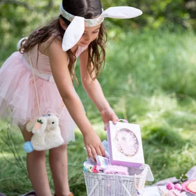 Mädchen im rosa Tutu-Kleid mit Hasenohren-Stirnband, draußen auf einer Wiese, trägt die Plüsch-Hasentasche umgehängt und packt einen Korb aus.