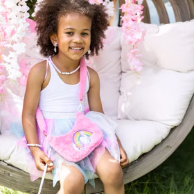 Einhorn Handtasche Rosa Plüsch Tasche Rosalie Regenbogenherz für Kinder Geschenkidee Geburtstag Kostüm-Zubehör Fasching Karneval Mottoparty Mädchen sitzt auf einer Schaukel mit Blütendeko, trägt ein Feenkostüm und die pinke Einhorn-Plüschtasche über der Schulter.