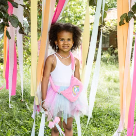 Einhorn Handtasche Rosa Plüsch Tasche Rosalie Regenbogenherz für Kinder Geschenkidee Geburtstag Kostüm-Zubehör Fasching Karneval Mottoparty Mädchen im bunten Tüllrock und Feenkostüm steht zwischen bunten Bändern im Garten, mit der pinken Einhorn-Handtasche umgehängt.