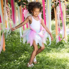 Einhorn Handtasche Rosa Plüsch Tasche Rosalie Regenbogenherz für Kinder Geschenkidee Geburtstag Kostüm-Zubehör Fasching Karneval Mottoparty Mädchen im weißen Oberteil und Tüllrock tanzt zwischen bunten Bändern im Garten, die pinke Einhorn-Tasche hängt quer über der Schulter.