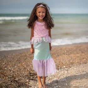 Mädchen trägt Meerjungfrauenkostüm und sitzt lächelnd auf einer alten Schatztruhe am Strand mit Meer im Hintergrund.