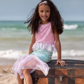 Meerjungfrauen Kostüm Mermaid Sirena Glitzerflosse für Kinder 3-8 Jahre Rosa Nixe Mermaid Kostüm Fasching Karneval Mottoparty Kindergeburtstag Mädchen trägt Meerjungfrauenkostüm und sitzt lächelnd auf einer alten Schatztruhe am Strand mit Meer im Hintergrund.