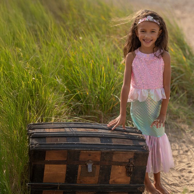 Mädchen trägt Meerjungfrauenkostüm und sitzt lächelnd auf einer alten Schatztruhe am Strand mit Meer im Hintergrund.