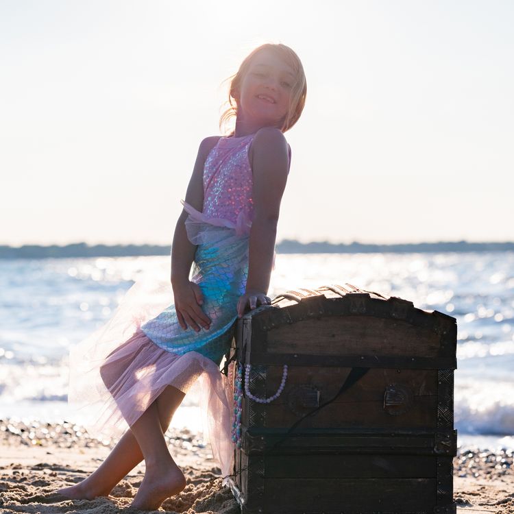 Mädchen trägt Meerjungfrauenkostüm und sitzt lächelnd auf einer alten Schatztruhe am Strand mit Meer im Hintergrund.