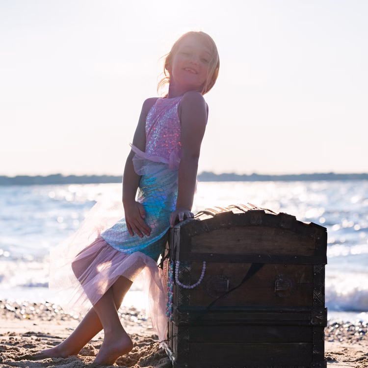 Meerjungfrauen Kostüm Mermaid Sirena Glitzerflosse für Kinder 3-8 Jahre Rosa Nixe Mermaid Kostüm Fasching Karneval Mottoparty Kindergeburtstag Mädchen im Meerjungfrauenkleid posiert bei Sonnenuntergang am Strand, lehnt sich an eine Schatztruhe.