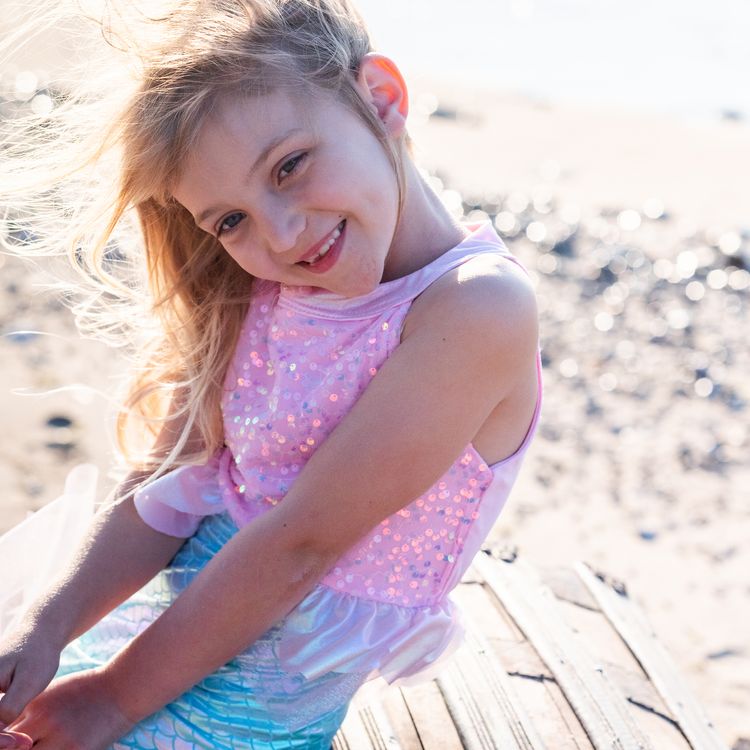 Mädchen trägt Meerjungfrauenkostüm und sitzt lächelnd auf einer alten Schatztruhe am Strand mit Meer im Hintergrund.