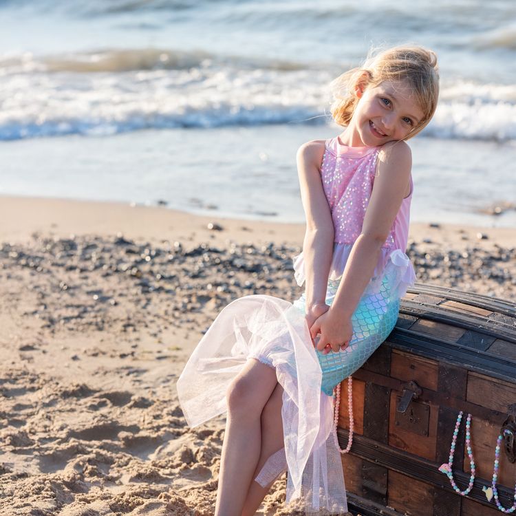 Mädchen trägt Meerjungfrauenkostüm und sitzt lächelnd auf einer alten Schatztruhe am Strand mit Meer im Hintergrund.