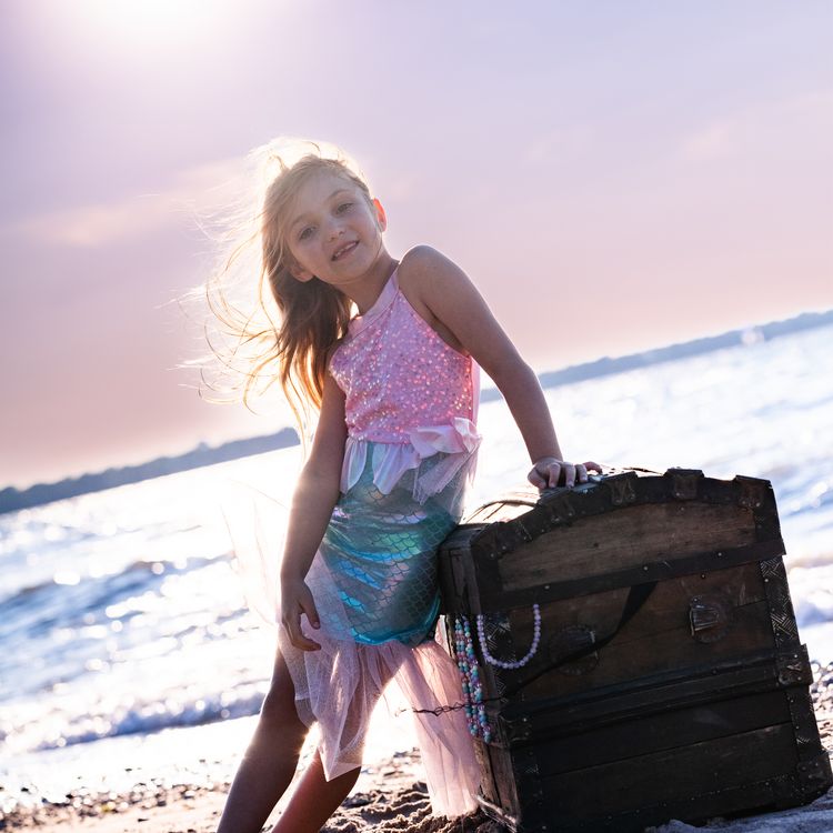 Mädchen trägt Meerjungfrauenkostüm und sitzt lächelnd auf einer alten Schatztruhe am Strand mit Meer im Hintergrund.