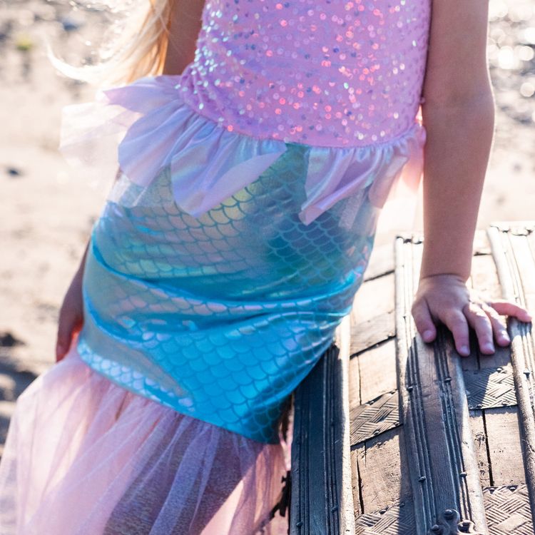 Mädchen trägt Meerjungfrauenkostüm und sitzt lächelnd auf einer alten Schatztruhe am Strand mit Meer im Hintergrund.