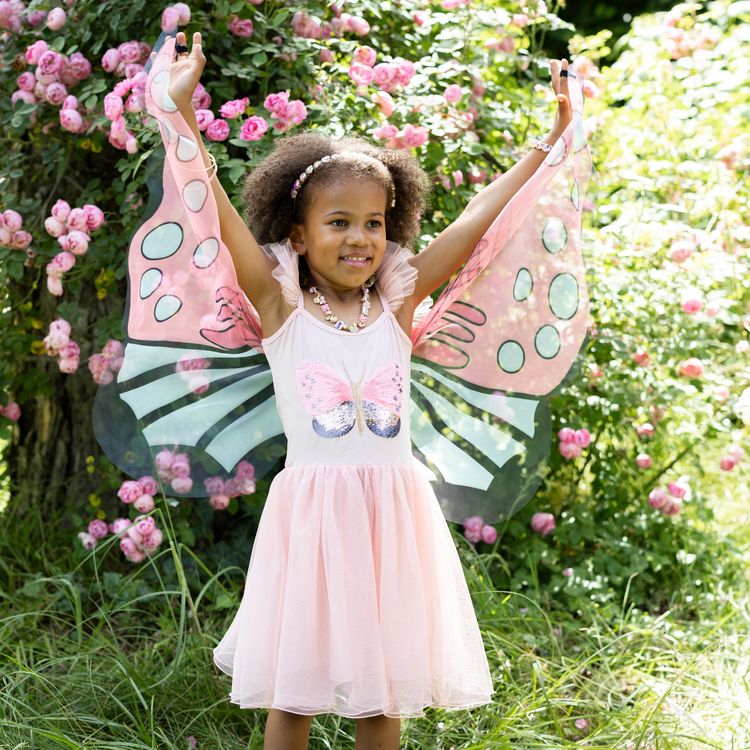 Schmetterling Kostüm Fee Rosalie Kleid mit Flügeln für Kinder Ein Mädchen im Schmetterlingskleid breitet die rosa Flügel im Garten vor einer Rosenhecke weit aus.