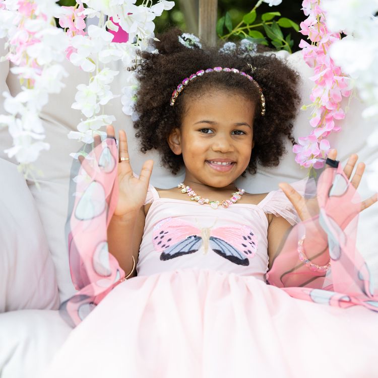 Schmetterling Kostüm Fee Rosalie Kleid mit Flügeln für Kinder Ein Mädchen im Schmetterlingskleid breitet die rosa Flügel im Garten vor einer Rosenhecke weit aus.