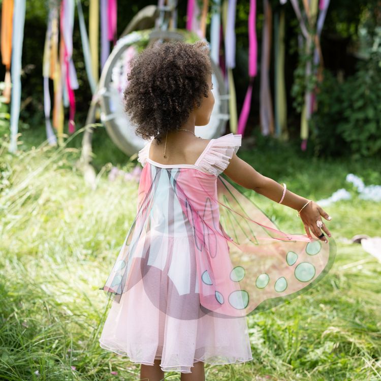 Schmetterling Kostüm Fee Rosalie Kleid mit Flügeln für Kinder Ein Mädchen im Schmetterlingskleid breitet die rosa Flügel im Garten vor einer Rosenhecke weit aus.