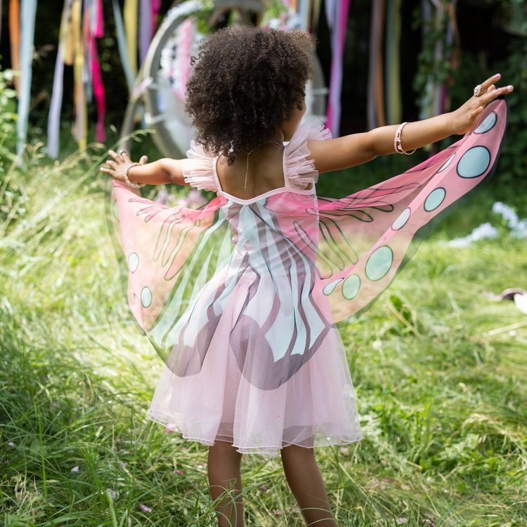 Schmetterling Kostüm Fee Rosalie Kleid mit Flügeln für Kinder Ein Mädchen im Schmetterlingskleid breitet die rosa Flügel im Garten vor einer Rosenhecke weit aus.
