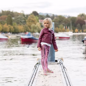 Leggings Rosa Metallic Fee Prinzessin Kostüm-Zubehör für Kinder 3-8 Jahre Kostüm-Zubehör Fasching Karneval Mottoparty Geburtstag Kinderfasching Ein Mädchen steht auf einem Steg am Wasser, trägt eine pinkfarbene Glanzleggings, eine weinrote Jacke und bunte Turnschuhe.