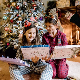 Zwei Mädchen in Weihnachts-Pyjamas sitzen vor dem Tannenbaum und betrachten ihre Adventskalender.