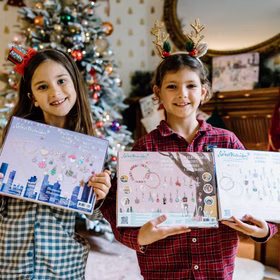 Zwei Kinder präsentieren stolz die Rückseiten der Adventskalender mit den Schmuckstücken vor einem geschmückten Weihnachtsbaum.