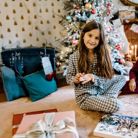 Mädchen im karierten Pyjama zeigt stolz die Armbänder aus dem Adventskalender, im Hintergrund ein geschmückter Weihnachtsbaum.