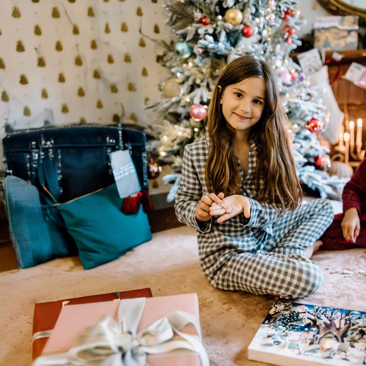 Schmuck Adventskalender 2 Armbänder & 22 Anhänger Cozy Christmas für Kinder Dezember Adventszeit Kalender für Kinder Weihnachten Mädchen im karierten Pyjama zeigt stolz die Armbänder aus dem Adventskalender, im Hintergrund ein geschmückter Weihnachtsbaum.