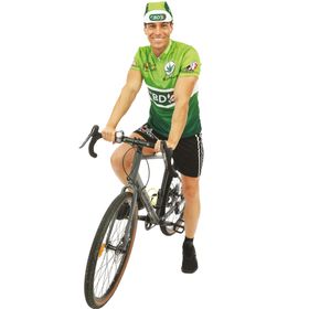 Mann im grünen Radfahrer-Kostüm mit Fahrrad, zeigt lachend auf sein Trikot.