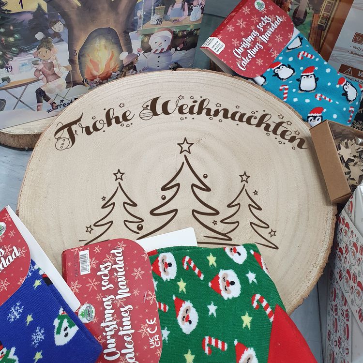 Große Baumscheibe mit Gravur, eingebettet in ein festliches Arrangement aus Tannenzweigen, goldenen Kugeln, Teelichtern und Kunstschnee, im Hintergrund warm leuchtende Bokeh-Lichter.