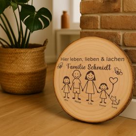 Personalisierte Holzscheibe mit Familienmotiv lehnt an einer Backsteinwand neben einer grünen Zimmerpflanze in einem natürlichen Wohnraumambiente.