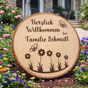 Personalisierte Baumscheibe mit Rinde und Gravur „Herzlich Willkommen bei Familie Schmidt“, dekoriert mit Schmetterlingen und Blumenmotiv, platziert in einem farbenfrohen Sommer-Blumenbeet am Hauseingang.