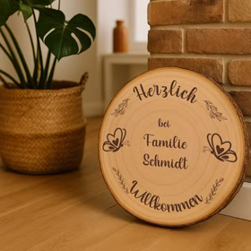 Personalisierte Holz-Baumscheibe mit Willkommensgravur als Dekoration im Flur, an eine Backsteinwand gelehnt neben Pflanze im Korb.
