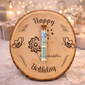 Baumscheibe mit „Happy Birthday“-Gravur und Wunscherfüller-Motiv, dekoriert mit einem mittig befestigten Reagenzglas mit Geldschein, stimmungsvoll arrangiert in festlicher Geburtstagsdekoration mit Kerzen und Geschenken.