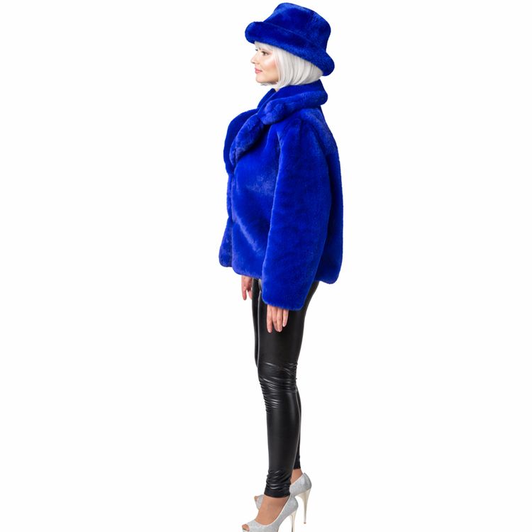 Dame trägt royalblaue Plüschjacke im 90er-Retro-Look mit passendem Plüschhut, schwarze Hose und silberne Pumps vor weißem Hintergrund.