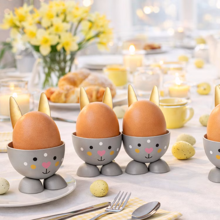 Eierbecher-Set 4 Stück Hase Gold aus Metall Osterhase gedeckter Tisch Ostern Oster-Deko Dekoration Frühling Osterfest Osterbrunch Tisch-Deko Mehrere graue Hasen-Eierbecher mit goldenen Ohren und braunen Hühnereiern auf einem hell gedeckten Oster-Frühstückstisch mit Blumen, Kerzen und Gebäck im Hintergrund.