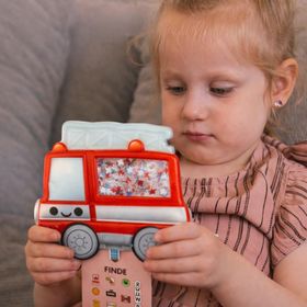 Sensorik-Spielzeug in Form eines roten Feuerwehrautos mit transparentem Fenster, gefüllt mit kleinen Perlen und versteckten Symbolen. Am unteren Rand hängt eine Suchkarte mit verschiedenen Feuerwehr-Motiven und Buchstaben.