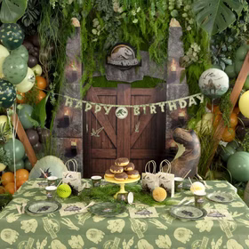 Ein fertig dekorierter Kindergeburtstagstisch im Jurassic World Stil mit Tischdecke, Geschirr und üppiger Dino-Dekoration.