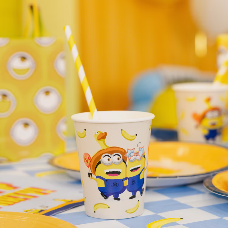 Minions Partybecher Tischdekoration Minions Partybecher auf blau karierter Tischdecke mit gelbem Teller, Bananenmotiven und farbenfroher Geburtstagsdekoration.