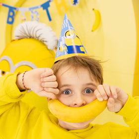 Minions Banana Party Kindergeburtstag Junge mit Minions Partyhütchen hält eine Banane wie ein Lächeln vor den Mund in einer leuchtend gelben Partydekoration.