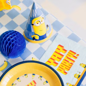 Minions Partyhut Tischdeko Yay Party Blaues Minions Partyhütchen neben gelbem Pappteller und Yay-Party-Serviette auf einer blau karierten Geburtstagstafel.