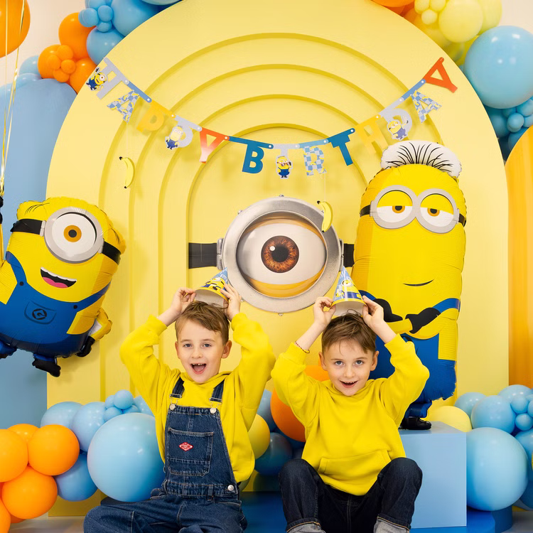 Minions Happy Birthday Party Kinder Zwei Jungen mit blauen Minions Partyhütchen vor Happy-Birthday-Banner, Ballons und großer gelber Minions Kulisse.