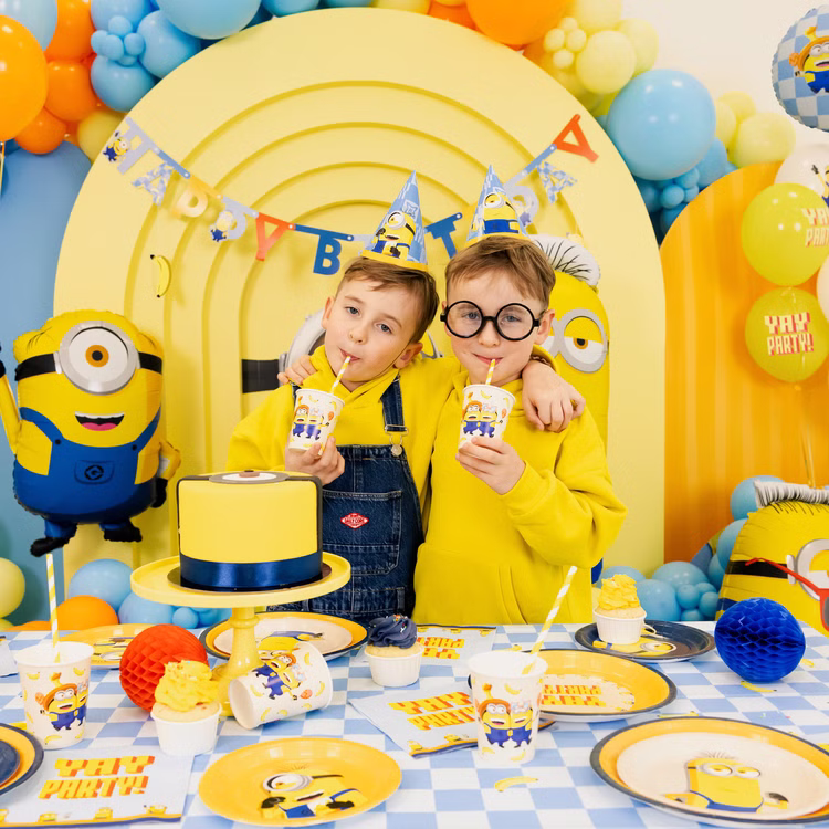 Minions Kindergeburtstag Partybecher Tisch Zwei Kinder trinken aus Minions Partybechern an einer komplett gedeckten Geburtstagstafel mit Tellern, Servietten und Partyhüten.