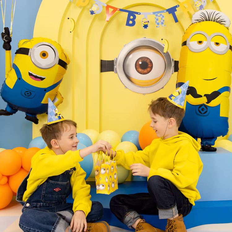 Minions Mitgebseltüte Kindergeburtstag Zwei Kinder mit Minions Partyhütchen überreichen sich eine gelbe Mitgebseltüte vor einer farbenfrohen Geburtstagskulisse.