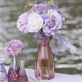 Rosa Glasvase Corolle mit pastellfarbenem Blumenstrauß und Schleife als dekoratives Arrangement am Wasser.